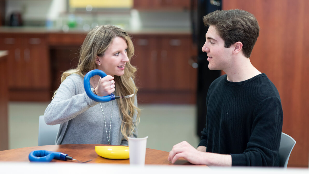 A professor demonstrates use of an accessibility-oriented kitchen utensil to an occupational therapy student.