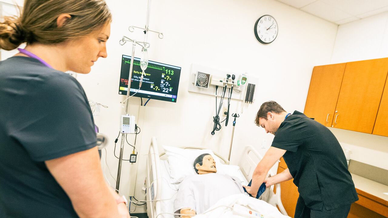 A male student puts a BP cuff on a mannikin while a female student observes