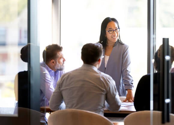 A woman standing and speaking at a conference table with 3 men sitting and listening to her