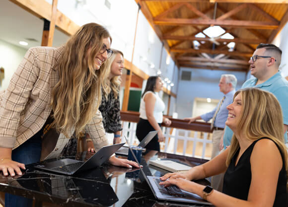 Two women with laptops smiling and talking in a bright workspace.