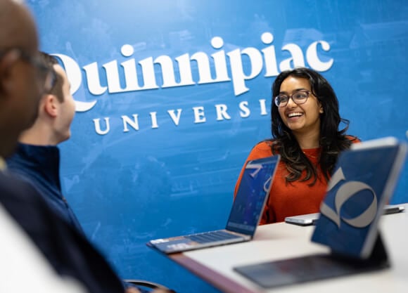 A female grad student sitting at a table with her laptop open smiling to someone off camera