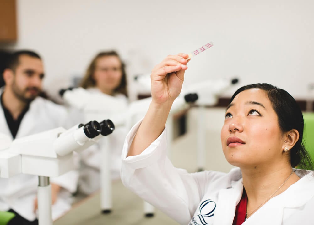 A student wearing a white lab coat holds a sample up to the light to inspect it