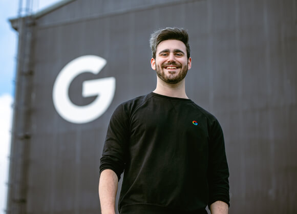 Quinnipiac alum Sam Bloch in a Google shirt smiles in front of a large Google G logo on a building