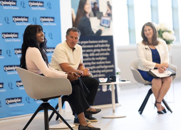 A graduate student speaks with a dean and trustee in front of a Quinnipiac backdrop