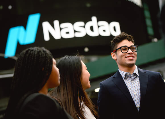Three Quinnipiac students in business suits stand in front of a Nasdaq sign