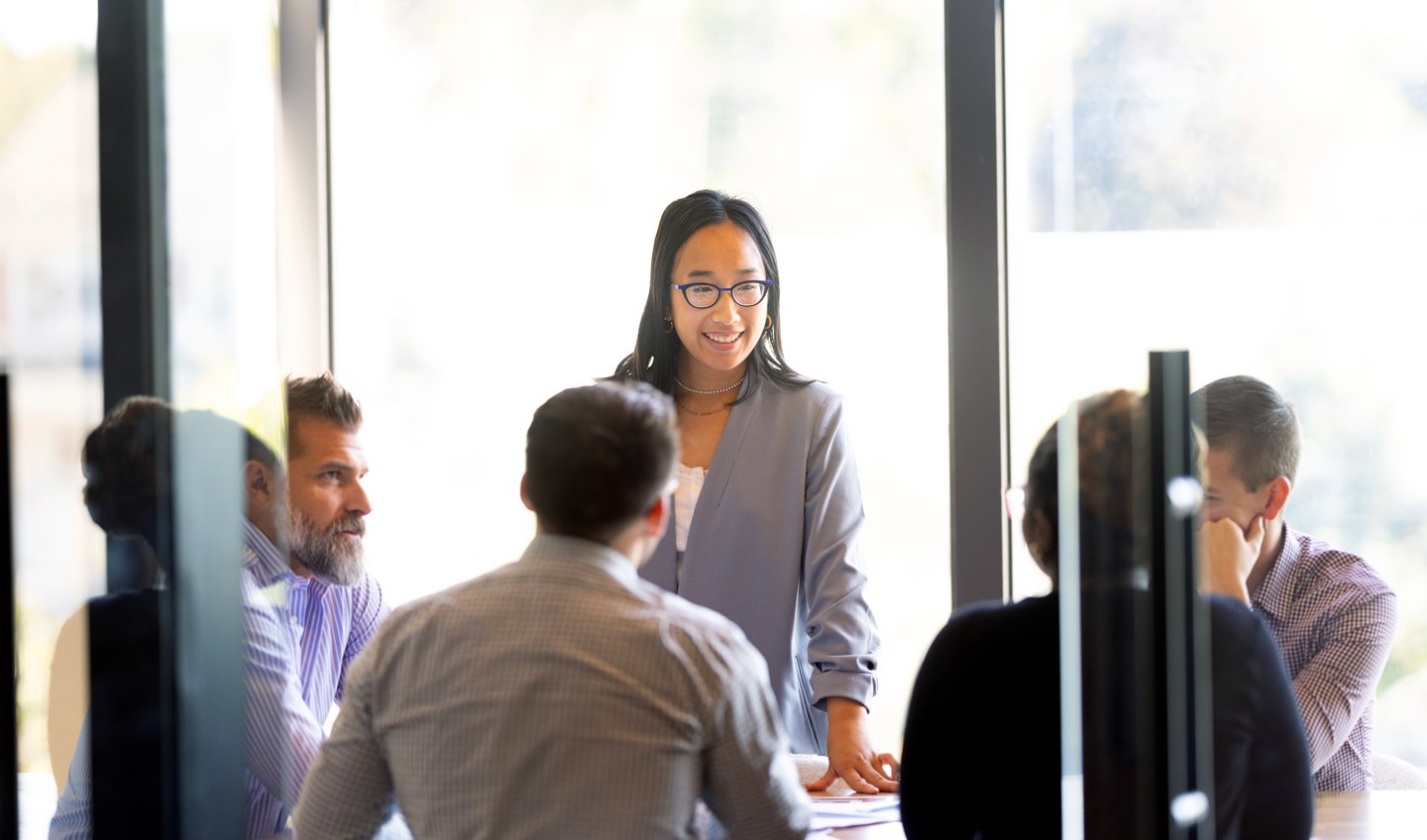 A graduate student stands at the head of a conference table presenting to a group of four peers