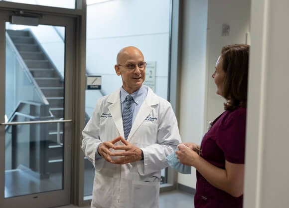 A male nursing professor in a lab coat talks to a woman wearing scrubs in an academic building hallway.